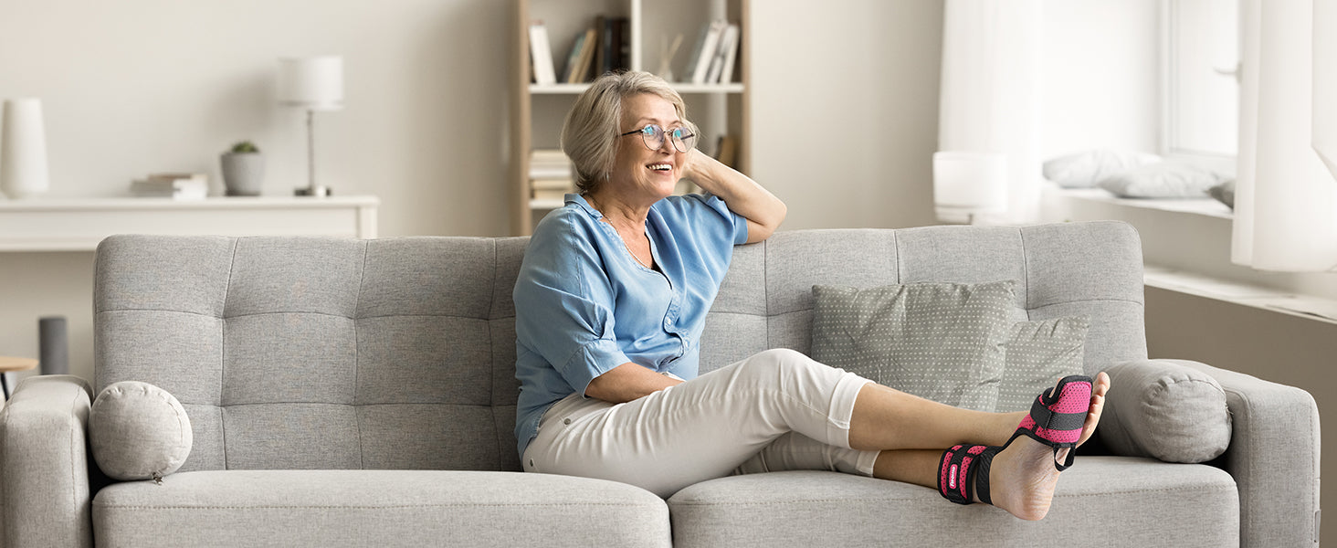 A smiling individual sitting comfortably on a couch, wearing a pink and black plantar fasciitis night splint on one foot over a white sock.