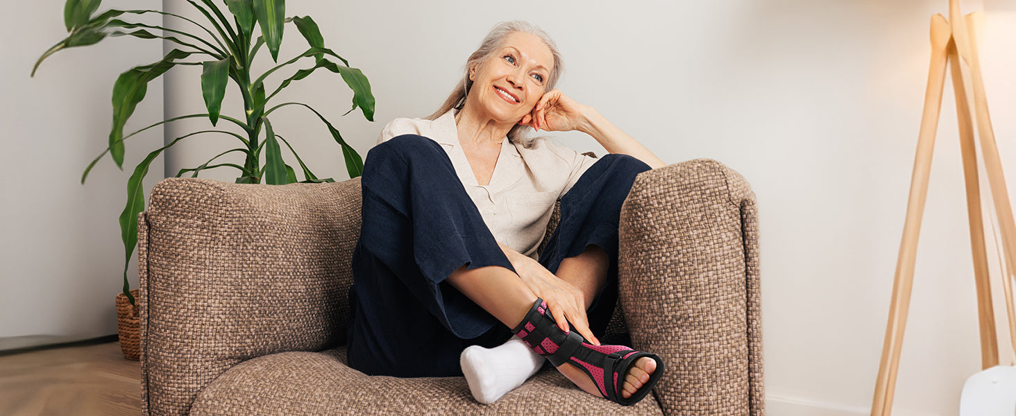 A person relaxing on a sofa in a living room, wearing a pink and black plantar fasciitis night splint on one foot, smiling with a hand resting on their head.