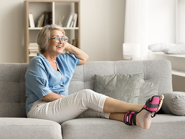 A smiling individual sitting comfortably on a couch, wearing a pink and black plantar fasciitis night splint on one foot over a white sock.